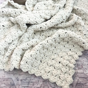 May include: A cream-colored, chunky crochet blanket featuring a repeating shell stitch pattern with a scalloped edge. The thick, textured yarn creates a soft, dense appearance throughout the fabric, which is folded to show its depth and volume.