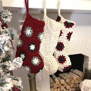 May include: Three handmade crochet Christmas stockings hanging from white hooks. One stocking is deep red with white and dark green floral motifs, while the other two are cream-colored with red and dark green accents, all featuring a hexagonal pattern.