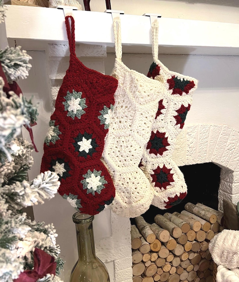 May include: Three handmade crochet Christmas stockings hanging from white hooks. One stocking is deep red with white and dark green floral motifs, while the other two are cream-colored with red and dark green accents, all featuring a hexagonal pattern.