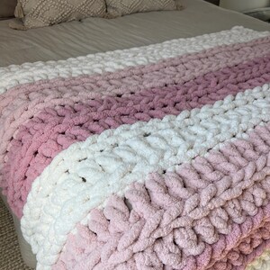 May include: A chunky knit throw blanket featuring horizontal stripes in alternating shades of cream, light pink, and dusty rose. The blanket is crafted from thick, soft yarn with a visible looped texture, creating a cozy and substantial appearance.