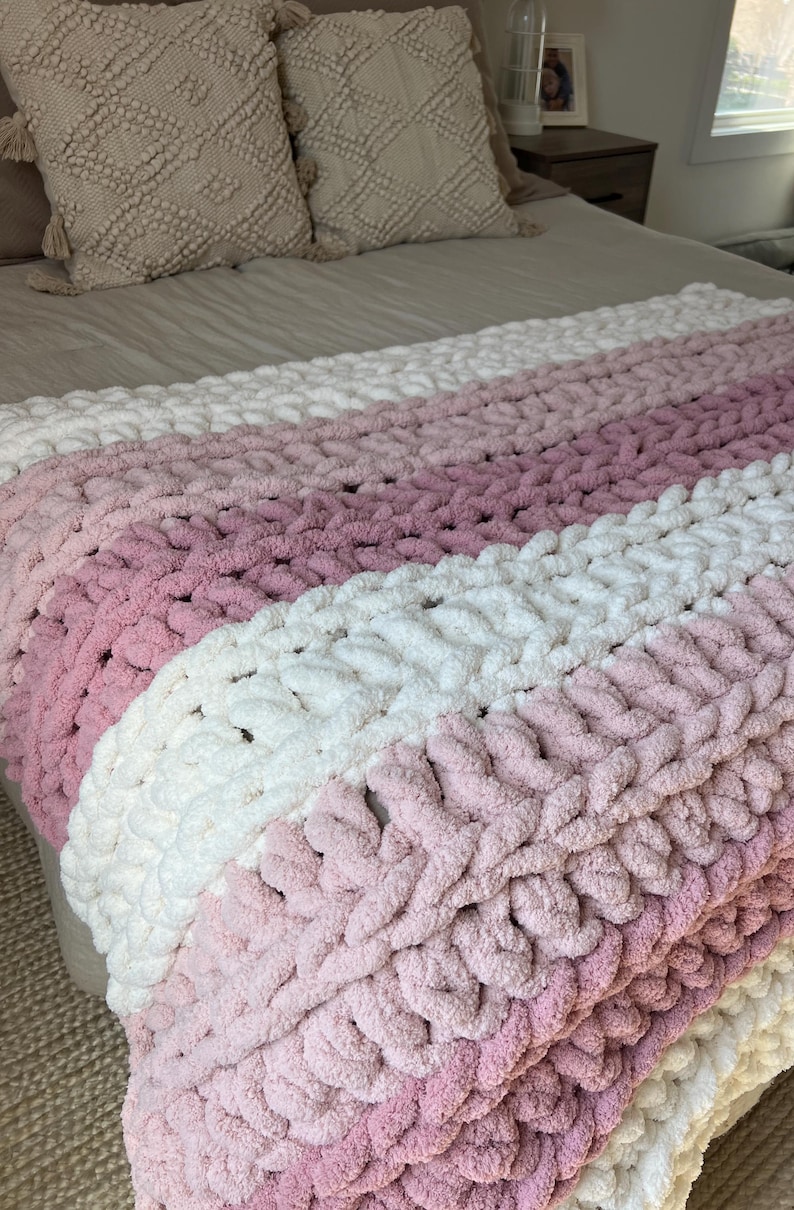 May include: A chunky knit throw blanket featuring horizontal stripes in alternating shades of cream, light pink, and dusty rose. The blanket is crafted from thick, soft yarn with a visible looped texture, creating a cozy and substantial appearance.