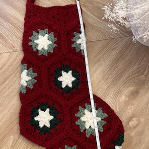 May include: A crocheted Christmas stocking made of deep red yarn with a hexagonal granny square pattern featuring cream and dark green centers. A white measuring tape placed along the side shows the stocking measures approximately 19 inches in length.
