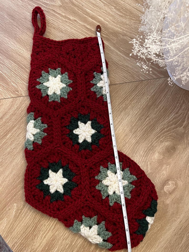 May include: A crocheted Christmas stocking made of deep red yarn with a hexagonal granny square pattern featuring cream and dark green centers. A white measuring tape placed along the side shows the stocking measures approximately 19 inches in length.