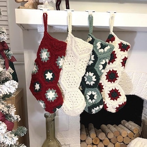 May include: Four handmade crochet Christmas stockings hanging in a row. The first is red with white and green floral motifs, the second is solid cream, the third is sage green with white motifs, and the fourth features a mix of red, green, and cream.