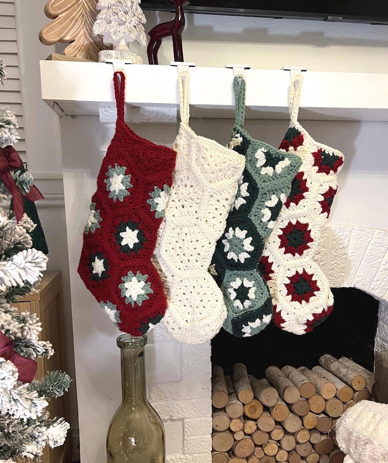 May include: Four handmade crochet Christmas stockings hanging in a row. The first is red with white and green floral motifs, the second is solid cream, the third is sage green with white motifs, and the fourth features a mix of red, green, and cream.