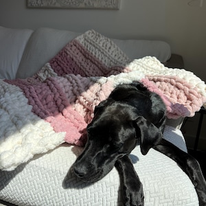 May include: A chunky knit throw blanket featuring horizontal stripes in alternating shades of cream, light pink, and dusty rose. The blanket is crafted from thick, soft yarn with a visible looped texture, draped over a textured white surface.