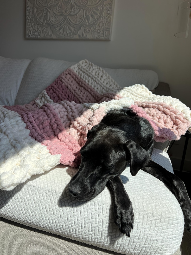 May include: A chunky knit throw blanket featuring horizontal stripes in alternating shades of cream, light pink, and dusty rose. The blanket is crafted from thick, soft yarn with a visible looped texture, draped over a textured white surface.