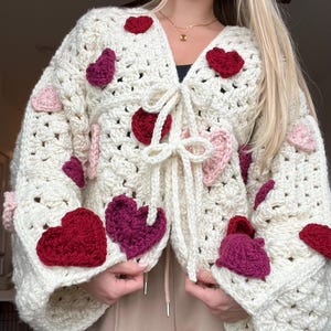 May include: A white crocheted cardigan with a tie front and large red, pink, and purple hearts.