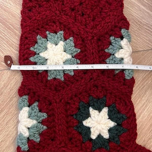 May include: A crochet Christmas stocking made of deep red yarn with integrated granny square motifs in cream, sage green, and dark forest green. A white measuring tape is placed across the stocking, showing a width of approximately 9 inches.