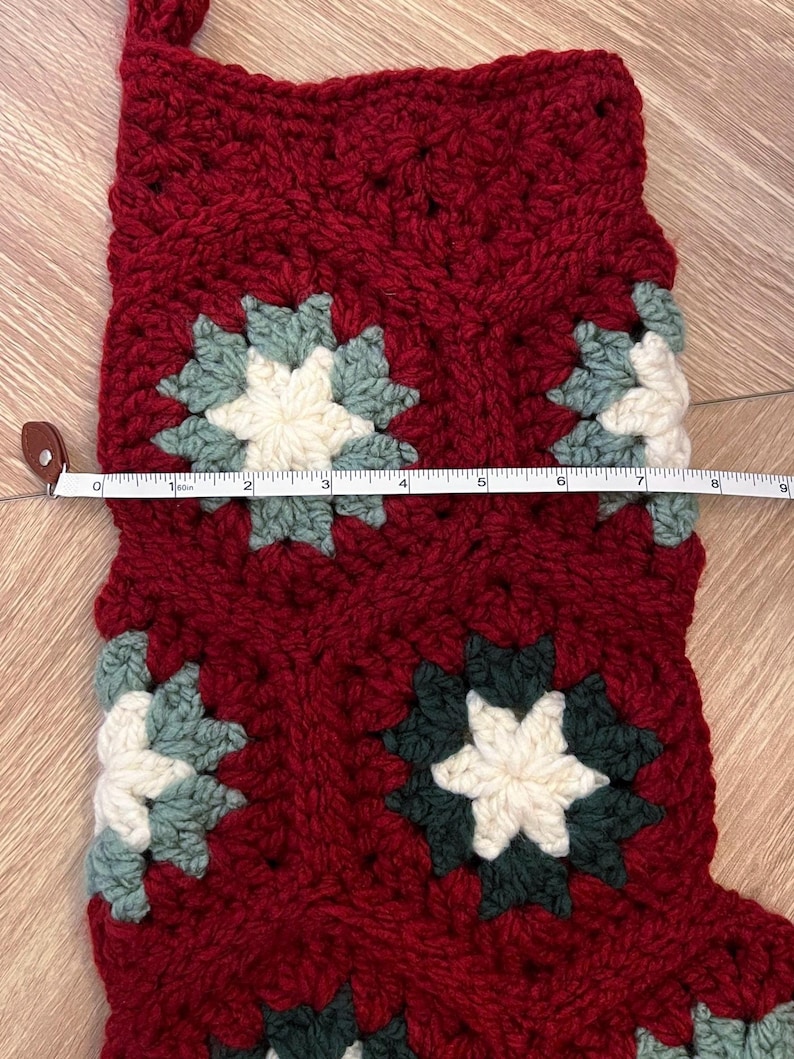 May include: A crochet Christmas stocking made of deep red yarn with integrated granny square motifs in cream, sage green, and dark forest green. A white measuring tape is placed across the stocking, showing a width of approximately 9 inches.
