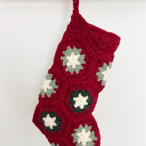 May include: A crocheted Christmas stocking made from deep red yarn, featuring a series of joined hexagonal motifs. Each hexagon contains a central cream-colored star shape surrounded by alternating dark green and sage green yarn accents.