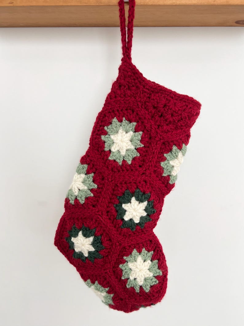 May include: A crocheted Christmas stocking made from deep red yarn, featuring a series of joined hexagonal motifs. Each hexagon contains a central cream-colored star shape surrounded by alternating dark green and sage green yarn accents.