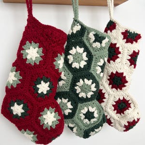 May include: Three handmade crochet stockings featuring a hexagonal granny square pattern. The first is primarily deep red, the second is sage and forest green, and the third is cream with red and green accents. Each has a matching yarn hanging loop.