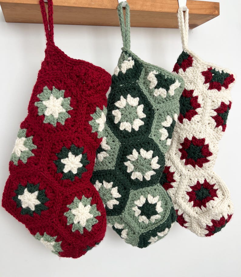 May include: Three handmade crochet stockings featuring a hexagonal granny square pattern. The first is primarily deep red, the second is sage and forest green, and the third is cream with red and green accents. Each has a matching yarn hanging loop.