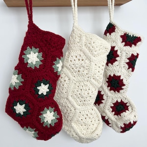 May include: Three crocheted Christmas stockings hanging in a row. The left stocking is deep red with white and dark green floral motifs. The center stocking is solid cream. The right stocking is cream with dark red and dark green floral motifs.