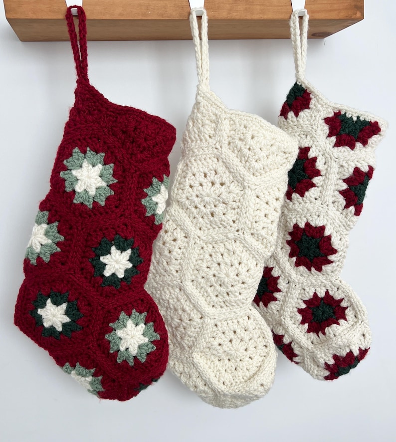 May include: Three crocheted Christmas stockings hanging in a row. The left stocking is deep red with white and dark green floral motifs. The center stocking is solid cream. The right stocking is cream with dark red and dark green floral motifs.