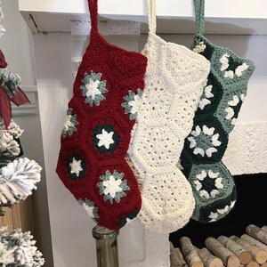 May include: Three handmade crochet Christmas stockings hanging in a row. The first is deep red with white and sage green floral motifs. The second is solid cream. The third is dark forest green with white and sage green floral accents.