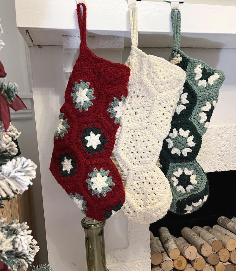 May include: Three handmade crochet Christmas stockings hanging in a row. The first is deep red with white and sage green floral motifs. The second is solid cream. The third is dark forest green with white and sage green floral accents.