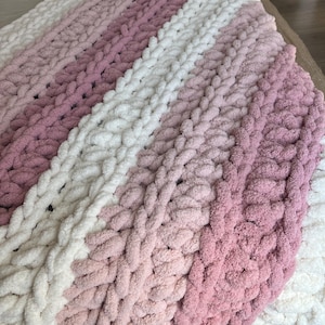 May include: A chunky knit throw blanket featuring thick, soft yarn in alternating horizontal stripes of cream, light pink, and dusty rose. The blanket is constructed with large, visible loops in a consistent, textured pattern throughout the fabric.