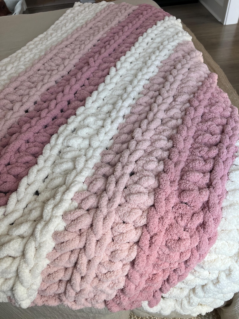 May include: A chunky knit throw blanket featuring thick, soft yarn in alternating horizontal stripes of cream, light pink, and dusty rose. The blanket is constructed with large, visible loops in a consistent, textured pattern throughout the fabric.
