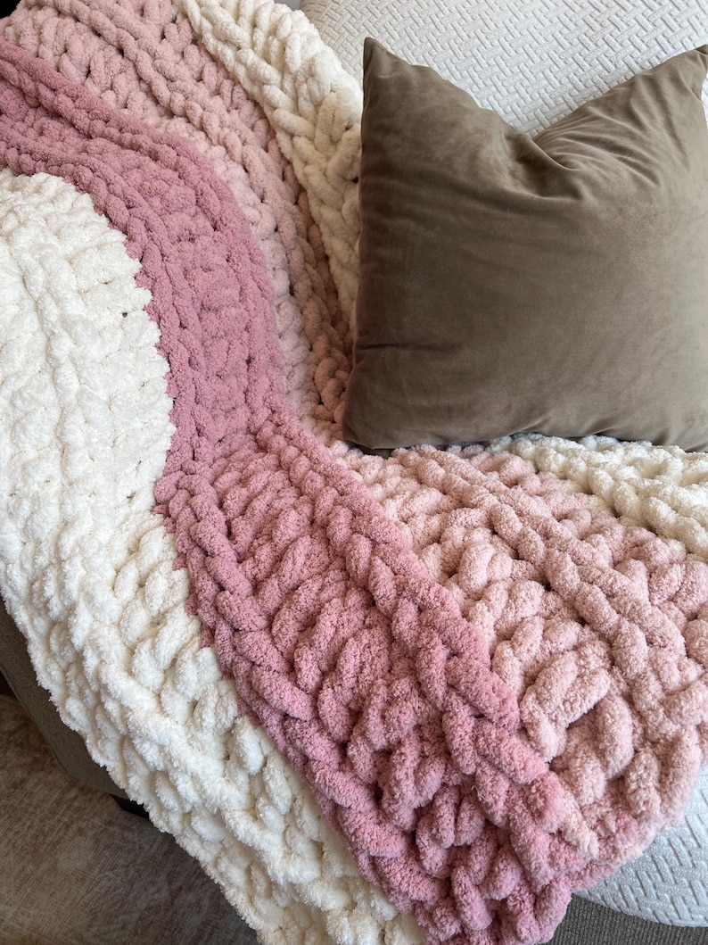 May include: A chunky knit throw blanket featuring thick, soft yarn in alternating diagonal stripes of cream, light pink, and dusty rose. A solid, smooth, taupe-colored square throw pillow rests on top of the textured, looped fabric of the blanket.