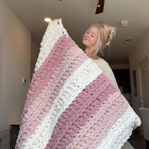 May include: A chunky knit blanket featuring thick, soft yarn in alternating horizontal stripes of cream white and dusty rose pink. The blanket is constructed with large, visible loops of plush, textured yarn, creating a thick and cozy appearance.