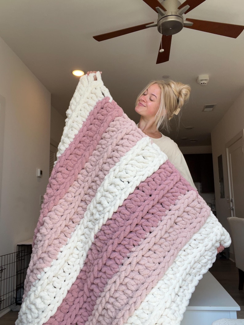 May include: A chunky knit blanket featuring thick, soft yarn in alternating horizontal stripes of cream white and dusty rose pink. The blanket is constructed with large, visible loops of plush, textured yarn, creating a thick and cozy appearance.