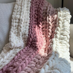 May include: A chunky knit throw blanket featuring thick, soft yarn in alternating vertical stripes of cream and dusty rose pink. The blanket has a highly textured, braided appearance created by large, interlocking loops of plush, fuzzy material.
