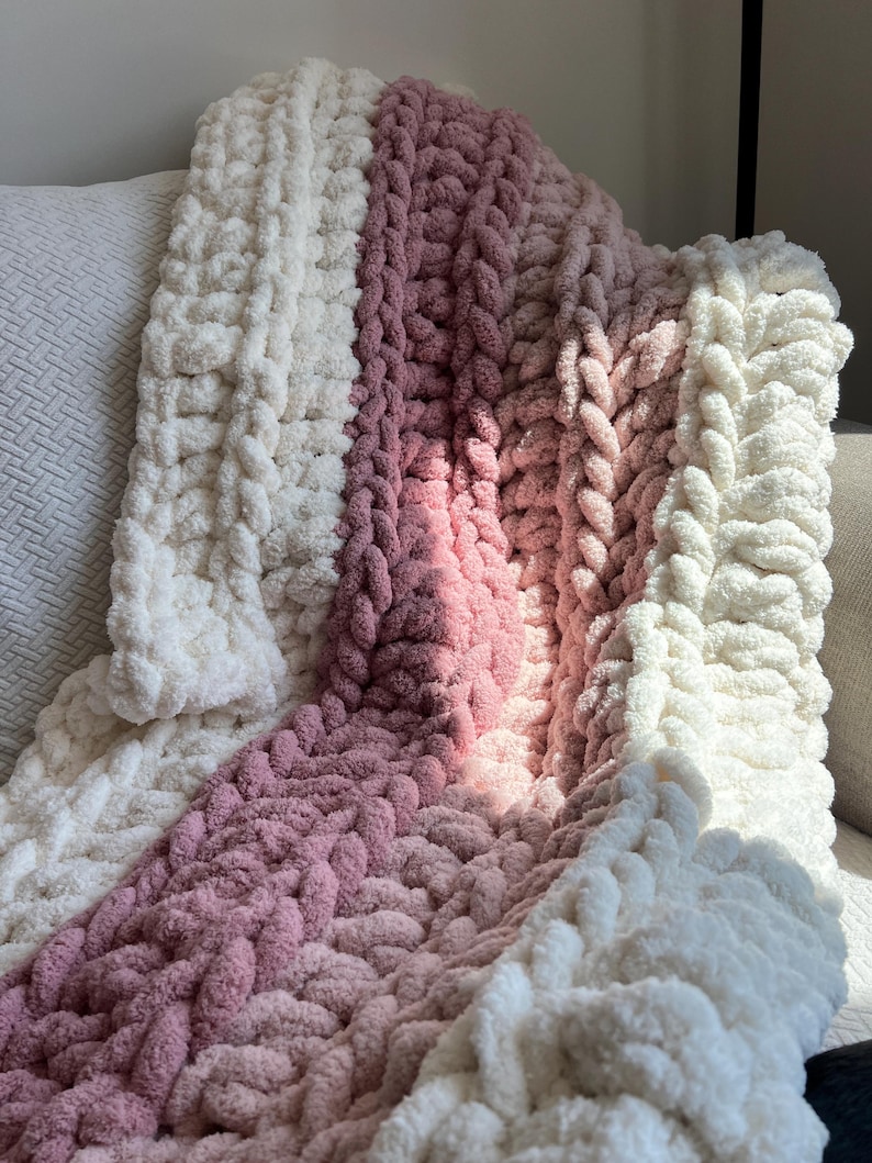 May include: A chunky knit throw blanket featuring thick, soft yarn in alternating vertical stripes of cream and dusty rose pink. The blanket has a highly textured, braided appearance created by large, interlocking loops of plush, fuzzy material.