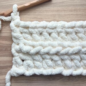 May include: A thick, white, chunky yarn crochet project in progress, featuring a textured cable stitch pattern. A large wooden crochet hook, marked as size 35/US 19.00mm, is inserted into the top loop of the soft, plush, white fiber textile.