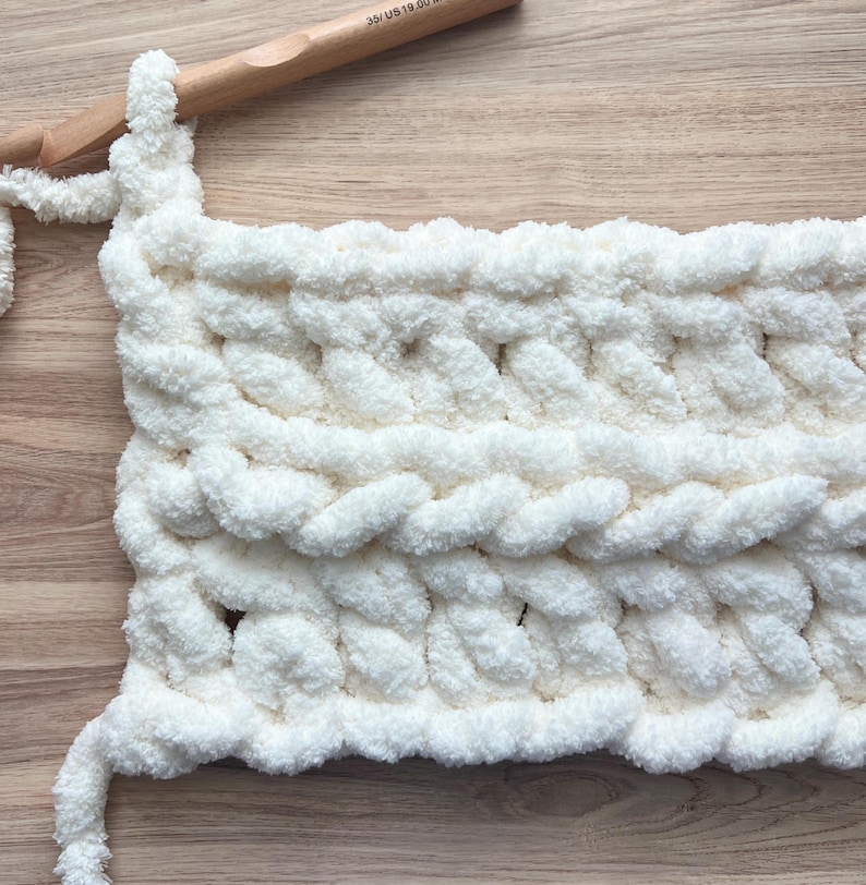 May include: A thick, white, chunky yarn crochet project in progress, featuring a textured cable stitch pattern. A large wooden crochet hook, marked as size 35/US 19.00mm, is inserted into the top loop of the soft, plush, white fiber textile.