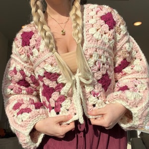 Peut inclure: Un cardigan crocheté fait à la main, de style granny square. Le vêtement présente un mélange de carrés roses, crème et bordeaux, avec une fermeture à nouer crème. Les manches sont longues et le style est décontracté et confortable.