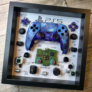 May include: A disassembled PlayStation 5 controller displayed in a black frame. The controller is painted with a galaxy design in shades of blue and purple. The frame includes labels for each part of the controller, including the direction pad, analog sticks, buttons, and vibration motors.