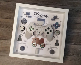 Peut inclure: Une présentation encadrée d'une manette de PlayStation One démontée. La manette est présentée dans ses composants, y compris les boutons, les joysticks analogiques et la carte de circuit imprimé. Le cadre est blanc et l'arrière-plan est en bois clair.