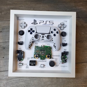May include: A white PlayStation 5 controller disassembled and framed. The controller is separated into its individual components, including the buttons, analog sticks, and vibration motors. The frame also includes the motherboard and other internal components. The text "PS5 INTRODUCED: 2020" is printed on the frame.