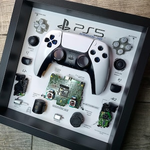 Op de afbeelding: Een ingelijste display met een gedemonteerde PlayStation 5-controller en zijn componenten. De witte en zwarte controller staat centraal, omringd door gelabelde onderdelen zoals het moederbord, knoppen en analoge sticks. De tekst "PS5 INTRODUCED: 2020" is zichtbaar.