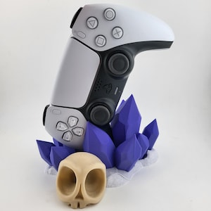 Può includere: Un controller PlayStation 5 bianco e nero è esposto su un supporto a forma di cristallo viola. Un piccolo teschio beige si trova nella parte anteriore del supporto.