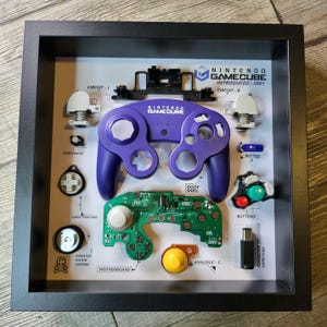 Puede incluir: Una vitrina enmarcada de un mando de Nintendo GameCube desmontado. El mando es morado y tiene una placa de circuito verde. El marco incluye etiquetas para cada parte del mando, incluyendo el botón de inicio/pausa, el pad direccional y la palanca analógica.