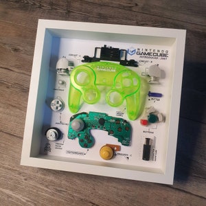 Puede incluir: Un mando de Nintendo GameCube desmontado en un marco blanco. El mando es verde y se ha separado en sus componentes individuales, incluyendo la carcasa del cuerpo, los botones, las placas de circuito y la palanca analógica. El marco incluye etiquetas que identifican cada pieza.
