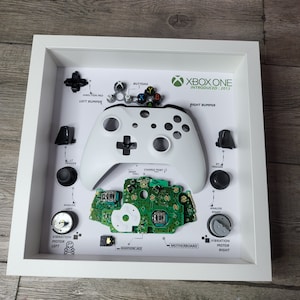 Puede incluir: Un mando de Xbox One blanco desmontado en un marco blanco. El mando está separado en sus partes individuales, incluidos los botones, los gatillos, las palancas analógicas y los motores de vibración. El marco también incluye etiquetas para cada parte, como "dirección pad", "bumper izquierdo" y "bumper derecho". El texto "XBOX ONE INTRODUCED: 2013" está impreso en la parte superior del marco.