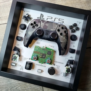 May include: A framed display featuring a PS5 controller with a camouflage design, disassembled components, and labeled parts. The frame includes the controller, motherboard, buttons, and vibration motors. The text "PS5 INTRODUCED: 2020" is visible.
