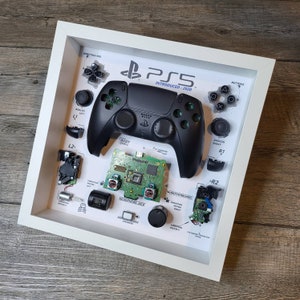 May include: A disassembled PlayStation 5 controller in a white frame. The controller is black with green accents and is labeled "PS5" and "Introduced: 2020". The frame also includes labels for each part of the controller, such as "Direction Pad", "Analog Left", "L2", "Body Shell", "Headphone Jack", "Vibration Motor Right", and "R2".