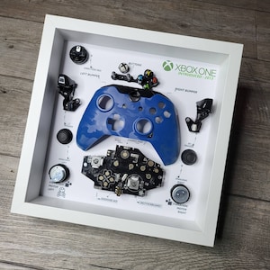 May include: A disassembled Xbox One controller with all of its parts laid out in a white frame. The controller is blue and has a camouflage pattern. The frame includes labels for each part of the controller, including the buttons, triggers, analog sticks, and vibration motors.
