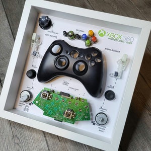 May include: A disassembled Xbox 360 controller displayed in a white frame. The controller is black and includes the body shell, buttons, triggers, analog sticks, and vibration motors. The frame also includes the text "XBOX 360 INTRODUCED: 2005".