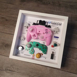 Puede incluir: Una exhibición enmarcada con componentes desmontados de un mando de Nintendo GameCube. El mando rosa es el punto focal, con otras piezas como botones y placas de circuito dispuestas alrededor. El texto incluye "NINTENDO GAMECUBE INTRODUCED: 2001".