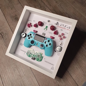 May include: A disassembled PlayStation 4 controller displayed in a white frame. The controller is teal blue with red buttons and a black faceplate. The frame includes a diagram of the controller's components, including the motherboard, buttons, and vibration motors. The text "PS4 INTRODUCED: 2013" is printed on the frame.