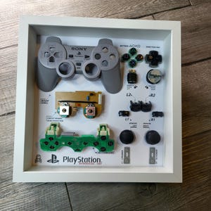 Puede incluir: Un mando de Sony PlayStation desmontado que se muestra en un marco blanco. El mando está separado en sus componentes individuales, incluyendo la carcasa, la placa de circuito, los botones y las palancas analógicas. El marco también incluye el texto "PlayStation" e "Introducido: 1997".