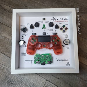 Puede incluir: Una presentación enmarcada de un mando de PlayStation 4 desmontado. El mando es rojo y transparente, y los componentes individuales están etiquetados con sus nombres. El marco es blanco y tiene un fondo negro.