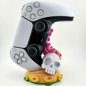 Pode incluir: Um controlador de videogame branco e preto está montado em um suporte impresso em 3D. O suporte é decorado com um polvo rosa, uma caveira branca e um design de recife de coral colorido.