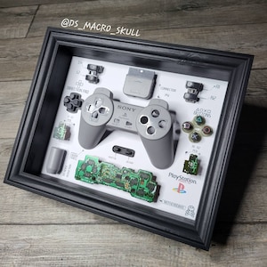 May include: A framed display of a disassembled PlayStation controller. The controller is gray and has a white background with the words "PlayStation" and "SONY" printed on it. The display also includes the controller's buttons, a memory card, and a circuit board.